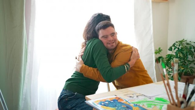 Young man with Down syndrome hugging his tutor when painting pictures indoors at home, homeschooling.