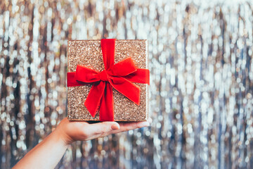 Female hand holding glittering golden gift box with velvet red ribbon with a bow on the sparkling gold tinsel background. The concept of a surprise gift for birthday, Xmas. Festive background.