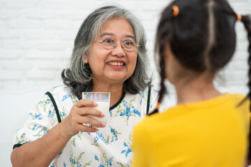 Happy elderly Asian Grandma sits beside her granddaughter and feeds fresh milk from glass for breakfast at home. Concept of a happy family and takes care together, preschool health care