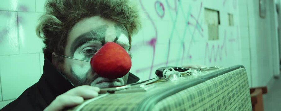 Portrait of sad clown with makeup and red nose sitting with suitcase in urban underpass and looking at camera