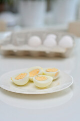 Boiled eggs on a kitchen table, healthy breakfast and protein rich diet. Eggs on a plate with a knife on a side, airy and white background with green plants and a light reflection from the table.