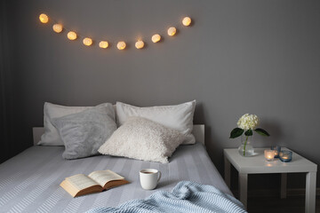 Stylish cosy bedroom in grey colors. Cozy interior, home decor. Bed with grey blanket, pillows, knitted plaid, bedside table, vase with hydrangea flower, candles, garland lights, book and cup of tea.