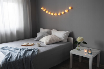 Stylish cosy bedroom in grey colors. Cozy interior, home decor. Bed with grey blanket, pillows, knitted plaid, bedside table, vase with hydrangea flower, candles, garland lights, book and cup of tea.