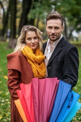 Obraz premium Beautiful couple in love stands in park holding a rainbow umbrella looking at the camera, Family couple autumn park in rainy weather