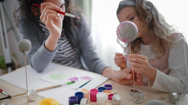 Little concentrated girl with her mother painting and making a solar system model at home, homeschooling.