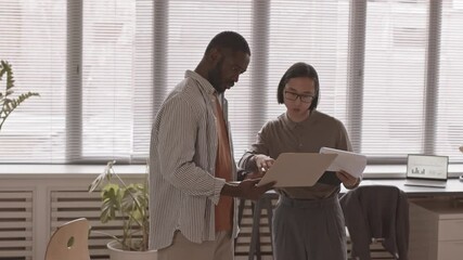 Medium slowmo shot of two multiethnic male colleagues talking while working on laptop standing by window in office