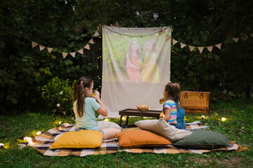 Open air cinema. Backyard Family outdoor movie night with kids. Sisters spending time together and watching cimema at backyard. DIY Screen with film. Summer outdoor weekend activities with children.