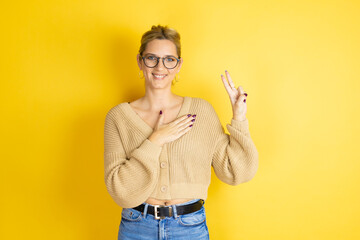 Young beautiful woman wearing casual sweater over isolated yellow background smiling swearing with hand on chest and fingers up, making a loyalty promise oath