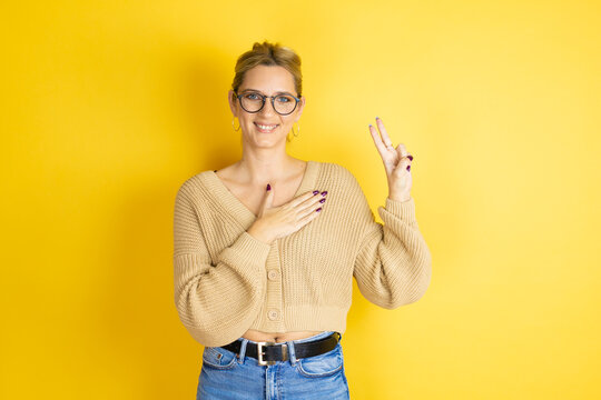 Young beautiful woman wearing casual sweater over isolated yellow background smiling swearing with hand on chest and fingers up, making a loyalty promise oath