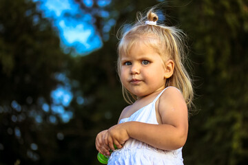 Naughty little girl in white dress space for text. Cute baby kid with blond hair looking seriously in green park. Small child daughter 2-4 years old. Cranky discontented bebe. Childhood. Bad mood kid.