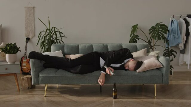 Young drunk guy sleeping on the couch in a suit wakes up in the morning with a hangover looking for a beer, drinks it and goes back to bed. Hard morning after night party with headache