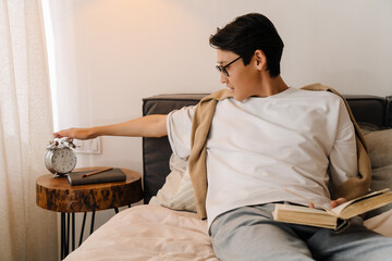 Asian boy in eyeglasses reading book while resting on bed