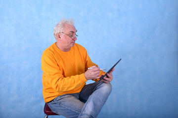 An elderly gray-haired man of retirement age draws with a stylus on a tablet computer on a blue...