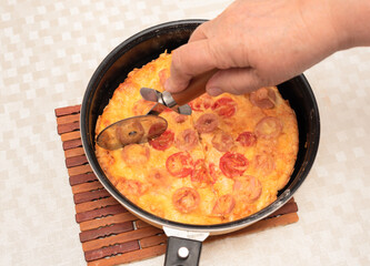 Ready to eat homemade pizza with sausages, tomatoes and cheese being cut with a pizza cutter by senior woman hand. Top view with selective focus. Process of cooking pizza, step 13