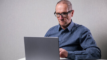 Senior man in classic dark blue dotted shirt with black-rimmed glasses works in computer sitting at white desk in office against grey wall.