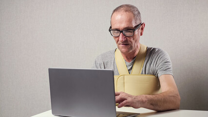 Senior man pensioner freelancer with glasses and fixating bandage over t-shirt works on laptop at...
