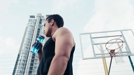low angle view of bi-racial athletic man holding sports bottle near basketball ring outdoors.