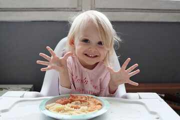 Cute little funny girl fooling around with food. A cheerful child is having lunch. A blonde-haired baby is eating at a table.