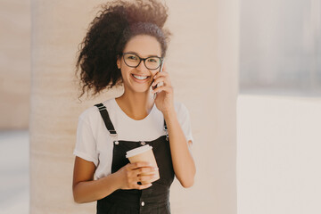 Pleasant looking curly teenage girl wears transparent glasses, talks on mobile phone, has coffee break, dressed in white t shirt and overalls, enjoys spare time and conversation. People and technology