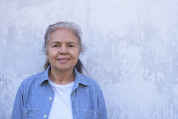 Front view medium to close up portrait of a happy senior Asian woman wearing blue shirt and white t-shirt, smiling, looking at camera, standing near old wall. Retirement and lifestyle concept