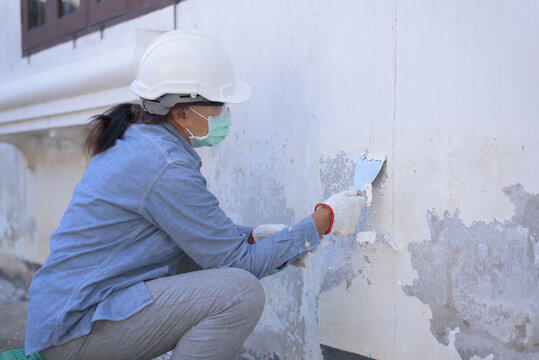 Side view, medium shot of senior Asian woman wearing safety hard hat, goggles, gloves and mask, holding trowel, sitting, peeling off paint film from old wall. DIY home renovation concept.