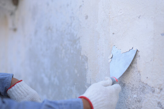 Close up, side view, selective focus with copy space of unrecognizable hand wearing gloves, holding trowel, peeling off paint film from old wall. Paint removing, preparing surface for painting concept