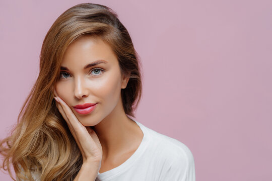 Close Up Shot Of Self Confident Beautiful Caucasian Woman Has Long Hair Combed On One Side, Touches Cheek With Palm, Wears Minimal Professional Makeup, Looks At Herself In Mirror, Prepares For Date