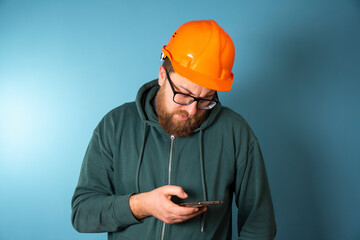 Cute Caucasian bearded construction worker with safety helmet on head and smartphone in hand. Engineer in orange helmet isolated on background. Construction worker concept .