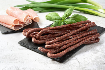 Hunting sausages on a light background. Raw smoked hunting sausages on a stone board with sliced pork balyk