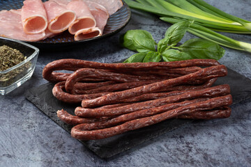 Hunting sausages on a dark background. Raw smoked hunting sausages on a stone board with sliced pork balyk