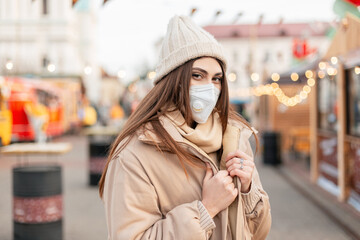 Stylish beautiful young girl model with a protective mask respirator in fashionable outerwear with a knitted hat, beige jacket and scarf walks in the city