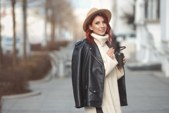 Urban fashion portrait of stylish millennial woman in modern clothes walking on city street with coffee