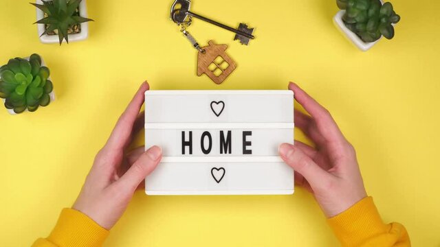 The hands of a young woman hold a white board with the inscription Home with hearts on a bright yellow background with green plants and apartment keys with a wooden keychain