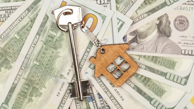 A silver key with a wooden keychain in the form of a house from new real estate is spinning against the background of many hundred dollar bills