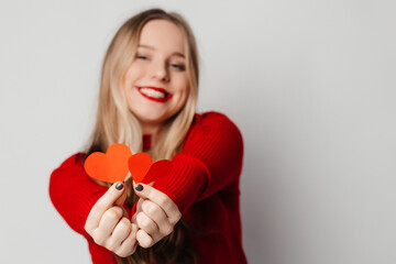Happy young blonde woman, dressed in red top giving red paper hearts to her boyfriend, standing against white background in a studio. Valentine's Day celebration concept