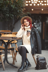 Full length street style portrait of fashionable and stylish redhead woman 25-30 years old in fashion casual clothes, sitting in cafe outdoors and drinking take away hot coffee from paper cup