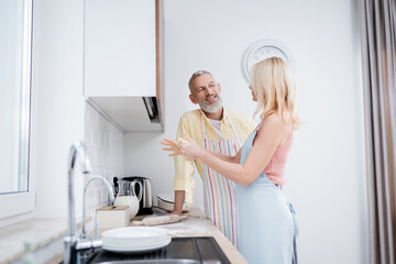 Positive mature man talking to wife with dough in kitchen.