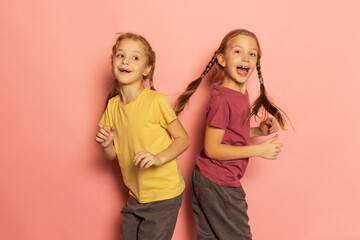 Cheerful little girls, sisters or siblings wearing casual style clothes dancing isolated on pink background. Concept of childhood, emotions