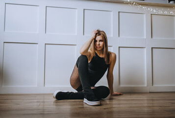 Beautiful girl model, sportswoman, blonde in black sportswear sits near a white wall in the interior.