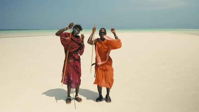 Two Young Happy Masai Tribe Members in Traditional Red Dresses and Sunglasses Dancing on the Sand Beach of the Indian Ocean. Zanzibar. Tanzania. 4K 50fps
