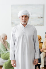 mature man in traditional muslim clothes looking at camera near blurred family.