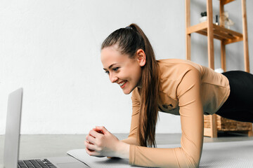 Close up of slim young girl with ponytail doing planking exercise indoors, looking at the laptop...