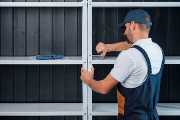 A uniformed worker is assembling a white metal shelving against a black clapboard wall.