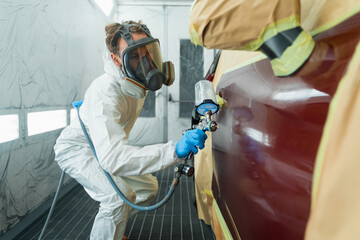 Auto painter in a protective suit and mask sprays a degreaser or other cleaner on a car door using a spray gun. Preparation of vehicle body elements for local repair