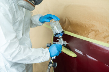 Close up view of auto painter in a protective suit and mask sprays a degreaser or other cleaner on a car door using a spray gun. Preparation of vehicle body elements for local repair