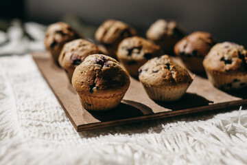 Homemade muffins with blueberries on brown wooden board on white table, boho napkin, leaves, sunlight. Tasty cakes, sweets, cupcakes. No sugar, free gluten. Receipt. Sweet dessert buffet. Mother’s day