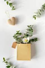 Craft bag with wintertime eucalyptus and white Helleborus flower. Brown paper tag and cardboard heart. Winter flat lay with evergreen twigs and winter rose. Copy-space on paper bag.