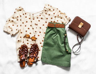 A set of women's summer clothes - bermuda shorts, polka dot blouse, braided leather sandals and a cross body bag on a light background, top view