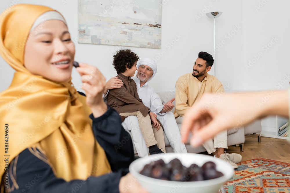 Wall mural blurred asian woman holding bowl with dates near interracial muslim family in living room.