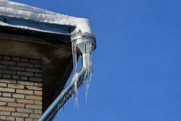 Bottom view of the corner of a snow-covered roof with a metal downpipe against the blue sky on a clear winter day. Large icicles hang from the drainpipe